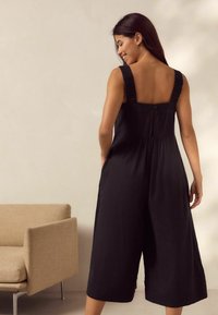Schwarzer Jumpsuit mit weit geschnittenen, kurzen Beinen, gerafften Schulterträgern und einem Knopfverschluss im Rücken. Glattes Material mit lockerer Passform.
