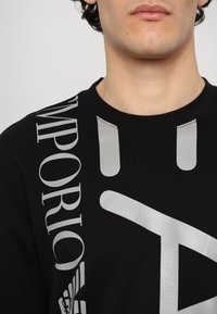 T-shirt nera in cotone con audaci grafiche e testo argentato, inclusa la scritta "EMPORIO", con un classico colletto a girocollo e una consistenza morbida.