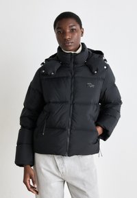 Giacca puffer nera con chiusura a zip, colletto alto e cappuccio rimovibile. Presenta un'imbottitura spessa, tasche laterali e un piccolo logo sul petto.