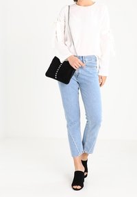 Biała bluzka z długim rękawem z ozdobnymi wstążkami, jasnoniebieskie, wysokoprofilowane dżinsy, czarna zamszowa torebka z srebrnymi ćwiekami oraz czarne mulesy z odkrytymi palcami.