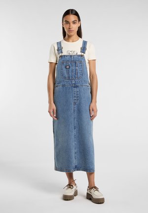 Vestito in denim azzurro in denim, con spalline regolabili, tasca frontale e spacco laterale. Abbinato a una camicia crema e scarpe con plateau beige.