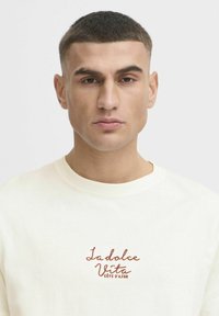 Lys kremfarget sweatshirt med teksturert stoff, ribbestrikket rund hals og brodert oransje "La dolce Vita Côte d'Azur" detalj på brystet.