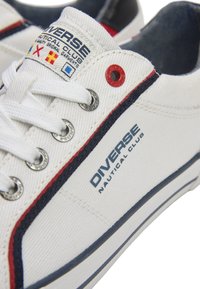 Białe płócienne trampki z niebieskimi i czerwonymi akcentami, metalowymi oczkami i tekstylną metką z napisem "Diverse Nautical Club."