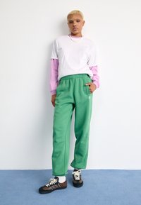 Henkilö, jolla on lyhyet blondit hiukset, seisoo päällään valkoinen t-paita vaaleanpunaisen pitkähihasen päällä, vihreät Adidas-housut, valkoiset sukat ja ruskeat lenkkarit.