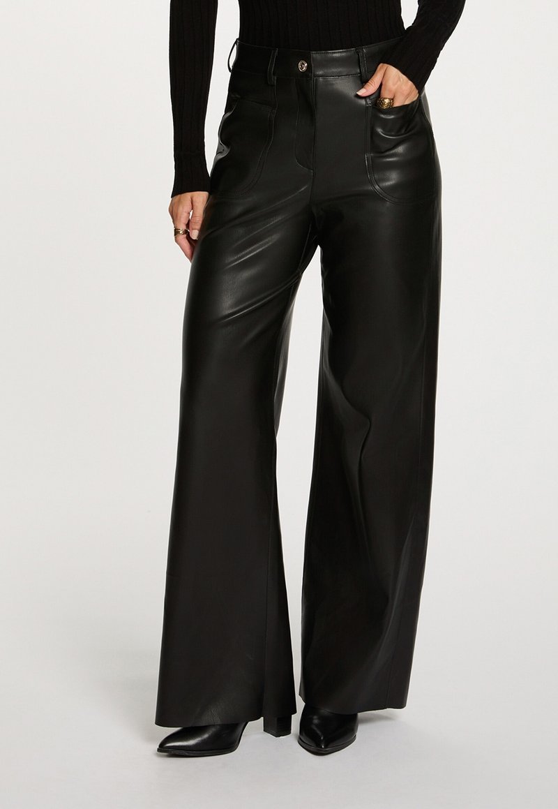 Personne portant un pantalon en cuir noir taille haute à jambes larges, des chaussures noires à bout pointu, et un haut noir à manches longues, main dans la poche.