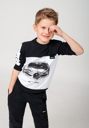 Maglietta a maniche lunghe bianca e nera con schizzo di un'auto e logo "BUGATTI", abbinata a pantaloni neri. Vestibilità giovanile, design casual.