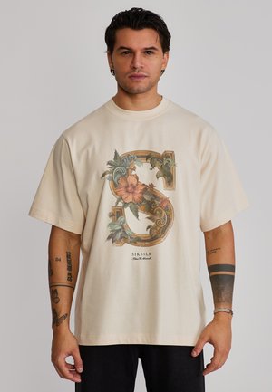 Uomo che indossa una T-shirt oversize color crema con grande motivo floreale ornamentale a forma di "S" e pantaloni neri, in piedi davanti a uno sfondo grigio chiaro.