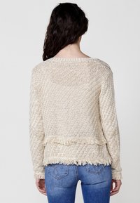 Beżowy dzianinowy sweter z teksturowanym wzorem jodełki, z frędzlowanym dołem i długimi rękawami. Widok od tyłu.