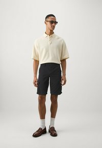 Polo shirt a maglia beige con maniche corte, abbinato a bermuda cargo scuri. Mocassini marroni con calze bianche completano l'outfit. Occhiali da sole arricchiscono il look.