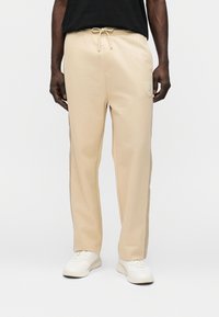 Μπεζ φόρμες με ελαστική μέση, κλείσιμο με κορδόνι, πλαϊνές τσέπες και υφασμάτινο λογότυπο στην αριστερή μηρό.