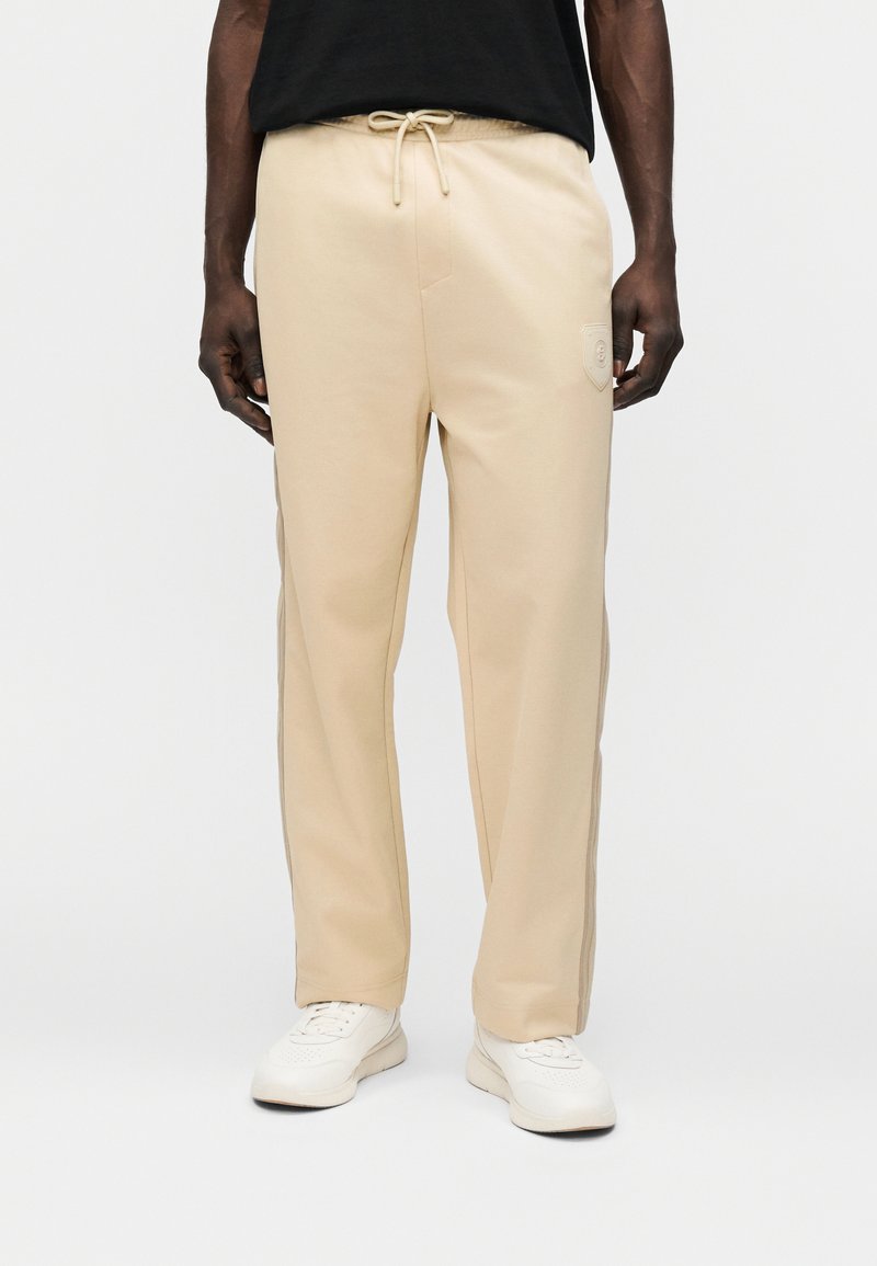 Μπεζ φόρμες με ελαστική μέση, κλείσιμο με κορδόνι, πλαϊνές τσέπες και υφασμάτινο λογότυπο στην αριστερή μηρό.