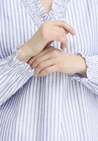 Blusa a righe blu e bianche con collo arricciato e polsini raccolti. Il tessuto sembra leggero e lavorato. Le mani sono posizionate vicino al collo.