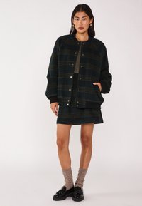 Giacca verde a quadri con polsini neri a coste, abbinata a una gonna coordinata. Il modello indossa mocassini neri e calze beige strutturate.