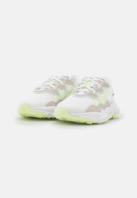 Zapatillas blancas con acentos verde claro, materiales de malla y ante, suela texturizada y detalles prominentes en los cordones. Forma redondeada con suelas gruesas.