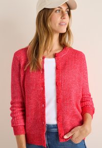 Cardigan rouge en tricot avec un col rond, fermeture à boutons, poignets côtelés et une surface texturée. Porté sur un top blanc et associé à du denim.