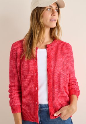 Röd stickad cardigan med rund halsringning, knappstängning, ribbade muddar och en strukturerad yta. Bärs över en vit topp och kombineras med denim.
