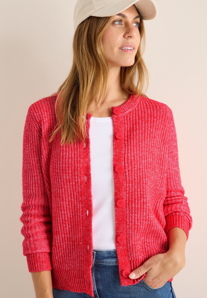 Cardigan rouge en tricot avec un col rond, fermeture à boutons, poignets côtelés et une surface texturée. Porté sur un top blanc et associé à du denim.