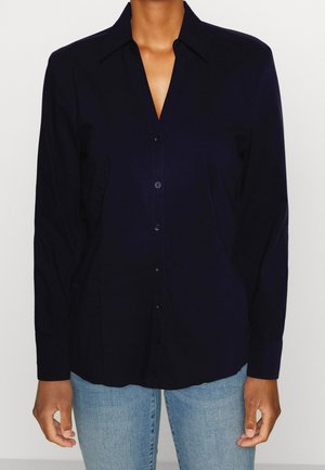 Person iført en langærmet sort skjorte med knapper og lyseblå jeans, stående med armene afslappet ned langs siden.