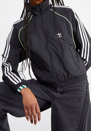 Zwarte Adidas trainingjas met witte strepen op de mouwen en een logo op de borst, gedragen met een donkere spijkerbroek en een groen-wit polsbandje.