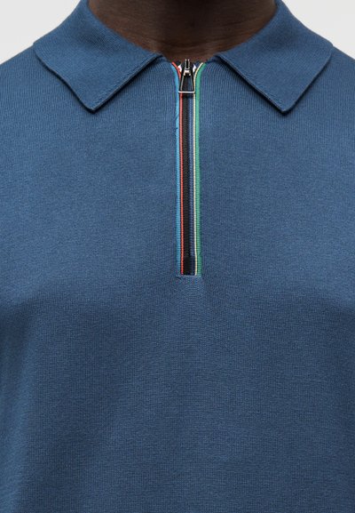 Άνδρας που φοράει μπλε πλεκτό πουκάμισο πόλο με πολύχρωμη ριγωτή φερμουάρ που τρέχει κάθετα στο γιακά.