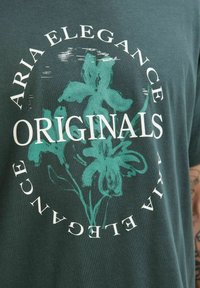 Mørkegrøn bomulds-T-shirt med et hvidt cirkulært grafisk design, der har teksten "ARIAL ELEGANCE ORIGINALS" og et teal blomsterdesign.