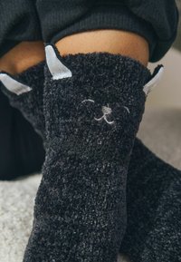 Svarta, fluffiga strumpor med en texturerad yta. Har gråa tygöron och broderade ansiktsdetaljer som liknar ett djurs ansikte.