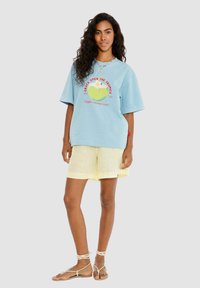 Světle modré oversize tričko s grafikou kokosu a textem, spárované se žlutými lněnými šortkami. Outfit doplňují sandály na pásky.