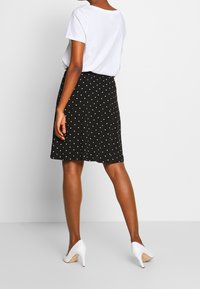 Femme portant un t-shirt blanc, une jupe noire longueur genoux à pois blancs, et des chaussures blanches à talons hauts, debout devant un arrière-plan uni.