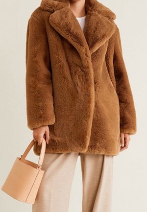 Brązowy płaszcz z futrzanym futerkiem noszony na jasnobeżowych spodniach, z torebką w stylu bucket w jasnobrązowym kolorze trzymaną w lewej ręce.