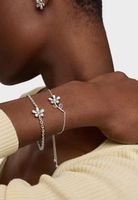 Bracciale in argento con catena legata, con un accento viola su un polso liscio, accanto a un tessuto lavorato a maglia testurizzato.