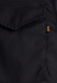 Pantaloni din material negru, cu o buzunar mare frontal, prevăzut cu clapetă și un mic eticheta cu logo portocalie. Materialul pare durabil și are o textură netedă.