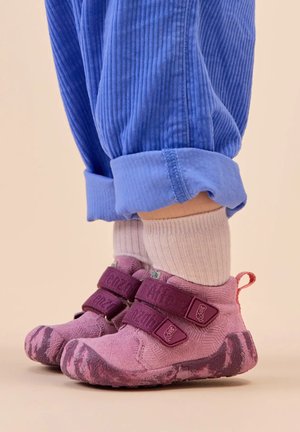 Pink, textured children's shoes with two purple Velcro straps and a patterned sole, paired with light pink socks and blue corduroy pants.