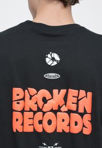 Μαύρο μπλουζάκι με έντονο, σπασμένο πορτοκαλί κείμενο "BROKEN RECORDS." Διαθέτει λευκό σπασμένο λογότυπο δίσκου πάνω από το κείμενο και λευκό γραφικό κάτω από αυτό.