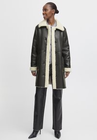 Olivgrüner Leder-Mantel mit krämigem Lammfellkragen und -manschetten, vorderen Knöpfen und seitlichen Taschen. Getragen über dunklen Hosen und Absätzen.