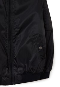 Zwarte pufferjas met een ritssluiting aan de voorkant, geribbelde manchetten en zijzakken met drukknoopsluiting. Glad, glanzend materiaal met een gewatteerd ontwerp.