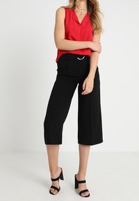 Punainen hihaton paita V-kaula-aukkolla; mustat leveälahkeiset housut; mustat korolliset sandaalit; housuissa yksinkertainen hopeinen koristenauha.