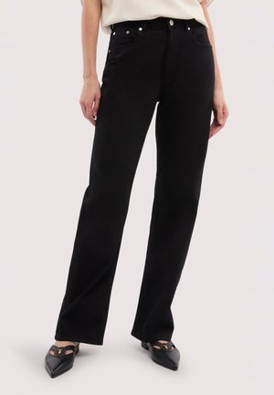 Person trägt schwarze Jeans mit geradem Bein, weißes Oberteil und schwarze spitze Schuhe mit goldenem Emblem, steht vor hellem Hintergrund.