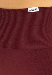 Leggings bordeaux avec une taille côtelée arborant l'étiquette "SMILODOX". Texture lisse, coupe ajustée et tissu extensible pour plus de confort.