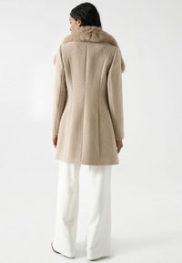 Manteau en laine beige avec un col en fourrure, coupe ajustée et coutures au dos. Associé à des pantalons blancs évasés et des chaussures noires minimalistes.