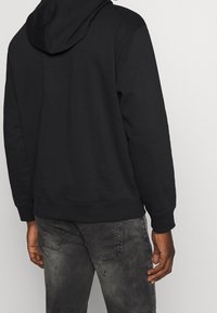 Muž v černé kapucíně a vybledlých černých džínách, zobrazený zezadu na jednoduchém světlém pozadí.