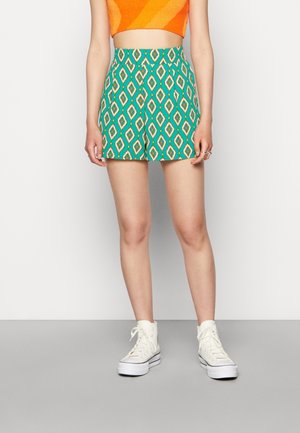 Personne portant un short vert taille haute à motif losange, un haut court orange et des baskets blanches montantes, debout devant un fond uni.