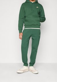 Person som bär en grön Nike-hoodie och matchande sweatpants med beige högskor, stående mot en enkel ljus bakgrund.