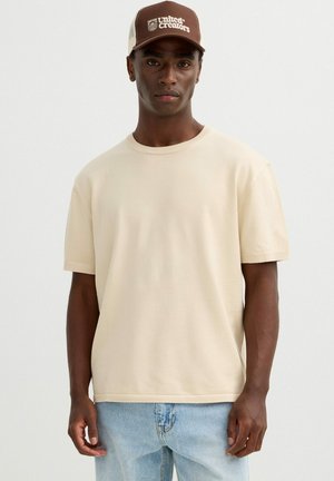 Hombre que lleva un suéter beige de manga corta, vaqueros azul claro y una gorra de béisbol marrón "United Creators", de pie frente a un fondo liso.