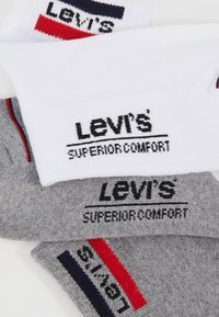 Trzy pary skarpetek Levi's: jedna biała z granatowymi i czerwonymi akcentami, jedna szara z czarnym napisem oraz czerwonymi i granatowymi paskami, z miękkiego materiału.