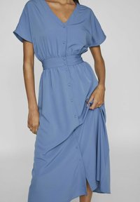 Sininen nappikauluspaita mekko, jossa on V-pääntie, lyhyet hihat, vyötärölle sidottu vyö ja laskostettu hame. Valmistettu pehmeästä kankaasta, jossa on laskeutuva rakenne.