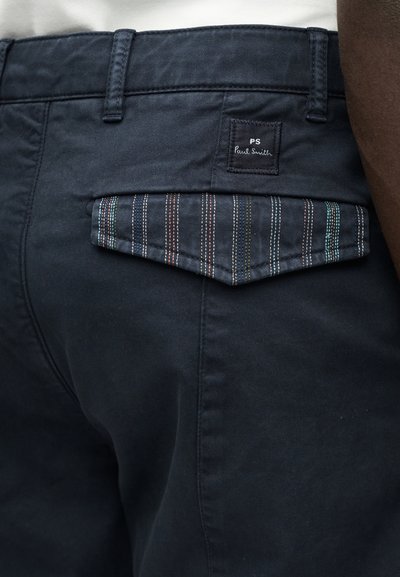 Κοντινό πλάνο σε ναυτικό παντελόνι με πίσω καπάκι τσέπης που διαθέτει πολύχρωμες ρίγες και ετικέτα "PS Paul Smith".