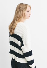 Jonge vrouw met steil blond haar kijkt naar rechts, gekleed in een wit en marineblauw geribbeld brei-sweater met brede horizontale strepen.