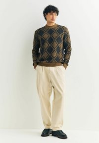 Uomo vestito con un maglione a diamante marrone e nero, pantaloni beige plissettati e scarpe nere, in piedi contro uno sfondo bianco con le mani in tasca.