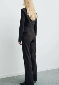 Completo nero su misura con giacca scolpita e pantaloni a gamba larga, realizzato in un tessuto liscio; visto da dietro, che espone capelli lunghi e lisci.
