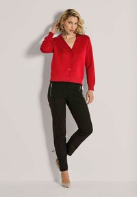 Frau mit kurzem blonden Haar, die eine rote strukturierte Knopfjacke, schwarze Hosen mit Reißverschlusstaschen und beigen High Heels trägt und posiert.